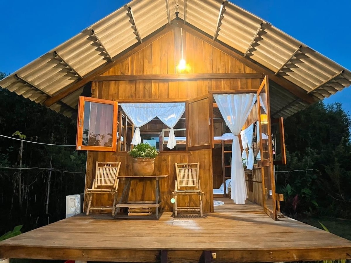 A wooden bungalow with large open windows allows for fresh air and natural light. A porch area is presented with two woven chairs and a potted plant, enhancing the connection to the surrounding nature. Soft lighting illuminates the entrance during twilight.
