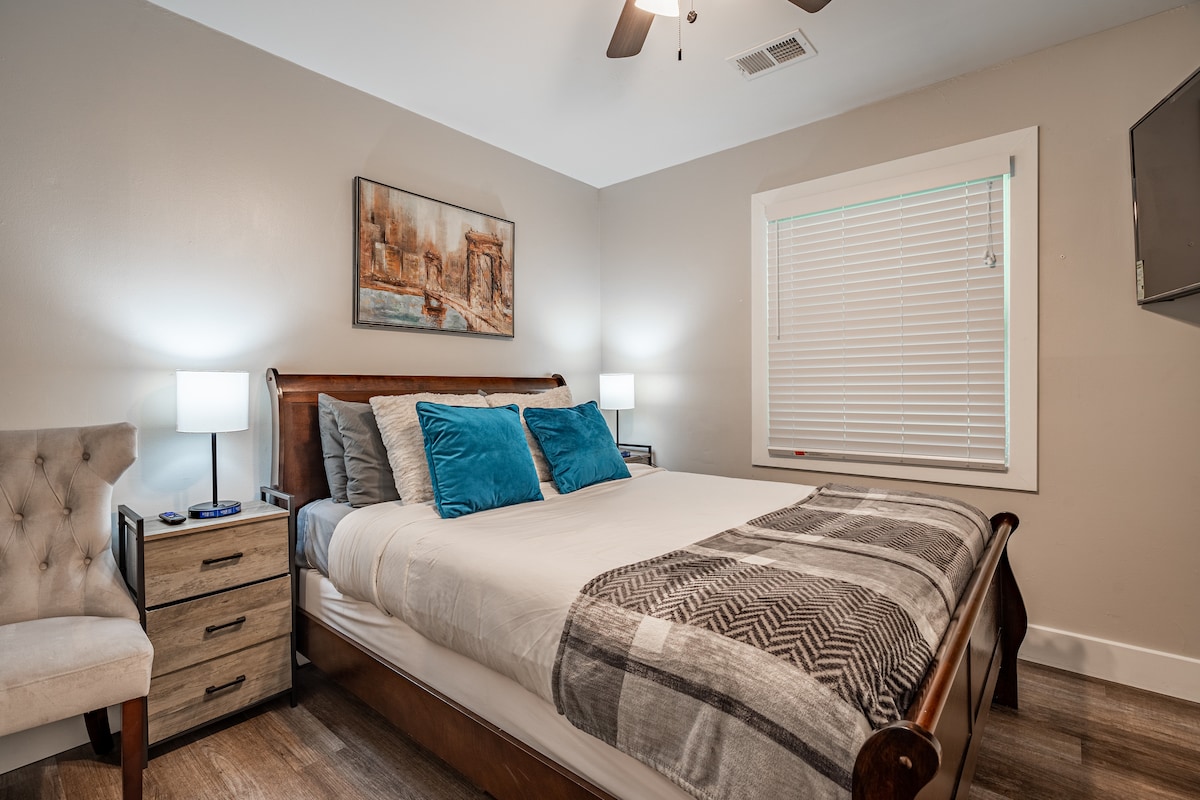 A comfortable bedroom features a spacious bed adorned with blue and gray pillows. Two lamps provide ambient lighting on each side, complementing the wooden nightstand. A window with white blinds allows natural light, while a piece of artwork enhances the neutral-toned walls. A plush armchair is situated nearby.
