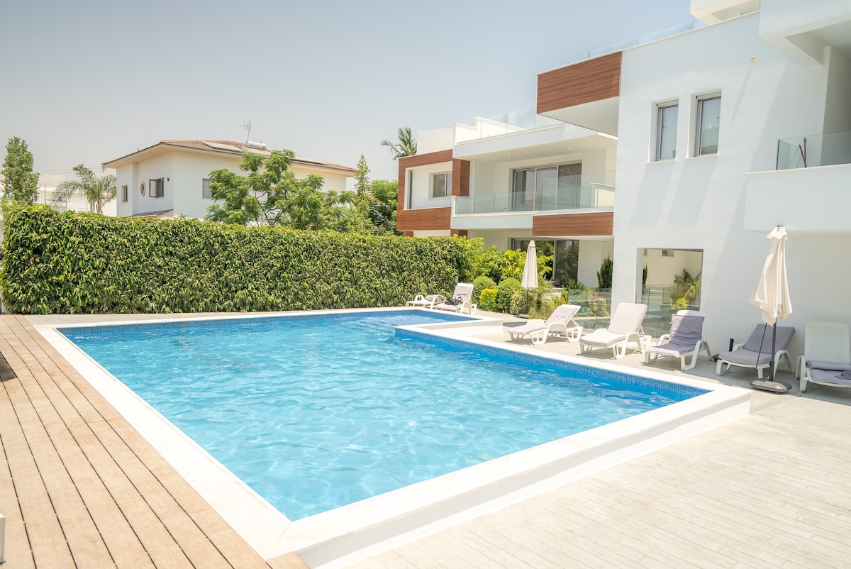 A large outdoor swimming pool is surrounded by a wooden deck and lounge chairs. Lush greenery provides privacy along the perimeter, while the bright blue water invites relaxation. The modern multi-story building in the background features balconies with glass railings.