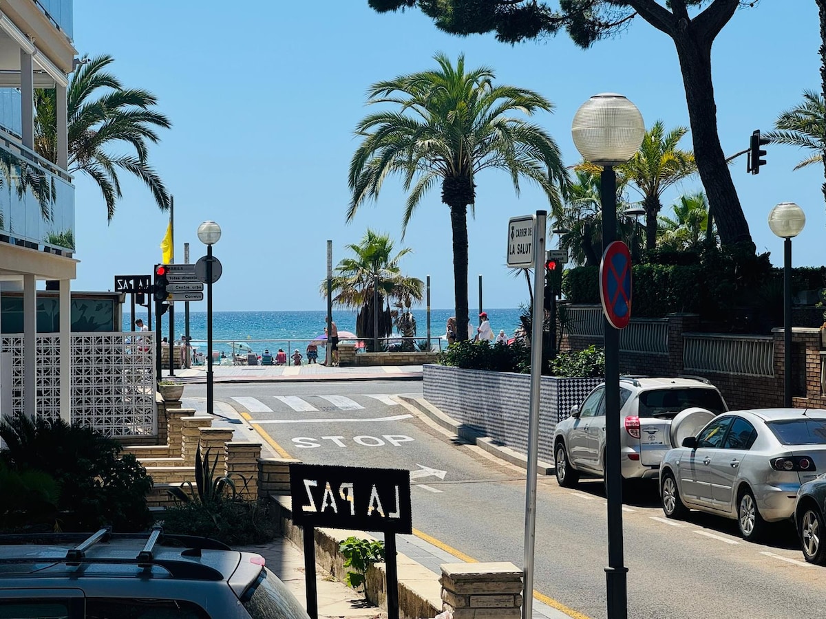 A view of the ocean is visible at the end of a palm-lined street. A crosswalk and a traffic sign are observed, with parked cars along the side. Sun loungers and umbrellas can be seen on the sandy beach, indicating accessibility to seaside leisure.