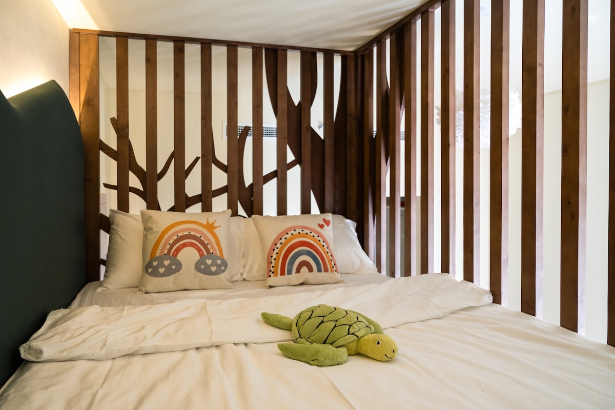 A cozy loft-style bed is framed by wooden slats resembling tree branches. Two colorful pillows with rainbow designs are placed on the spacious bed. A soft, green turtle plush toy adds a playful touch to the inviting sleeping area.