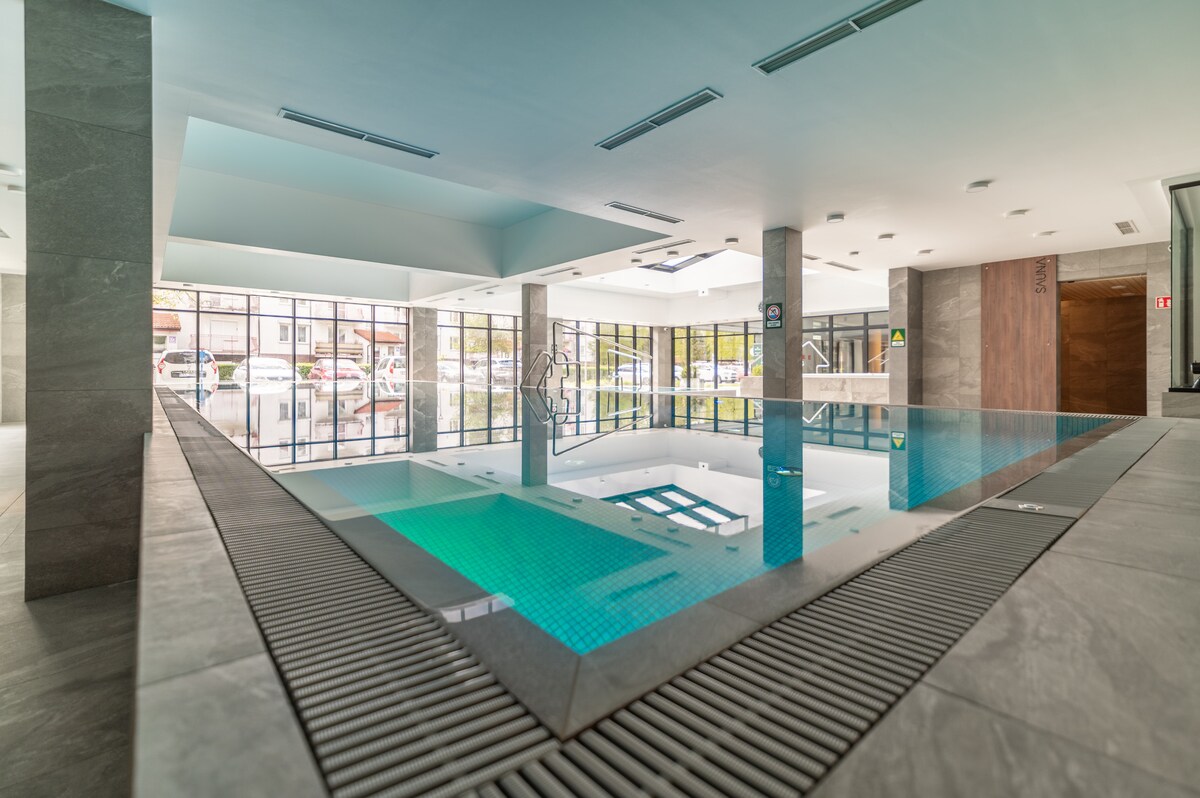 An indoor swimming pool with clear turquoise water is surrounded by large glass windows allowing natural light to fill the space. The sleek, modern design features gray stone walls and a smooth tile floor with a drainage strip along the perimeter.