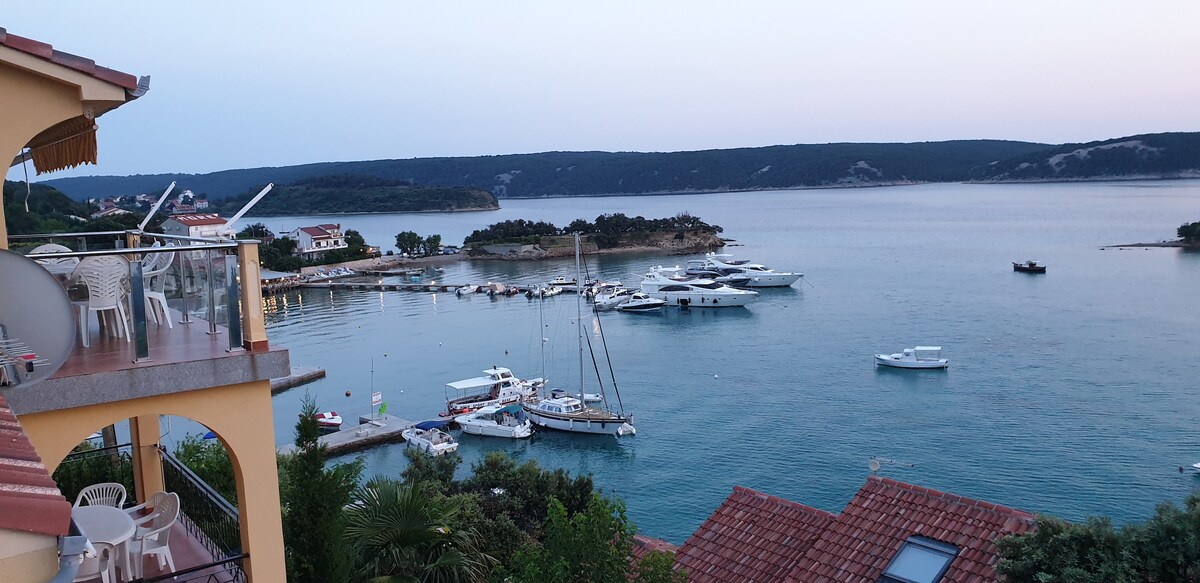 A scenic view of a tranquil bay, featuring anchored boats and a gentle shoreline. The horizon showcases a soft gradient of colors as the sun sets. Balcony seating is visible, allowing for enjoyment of the serene landscape and nearby marina.