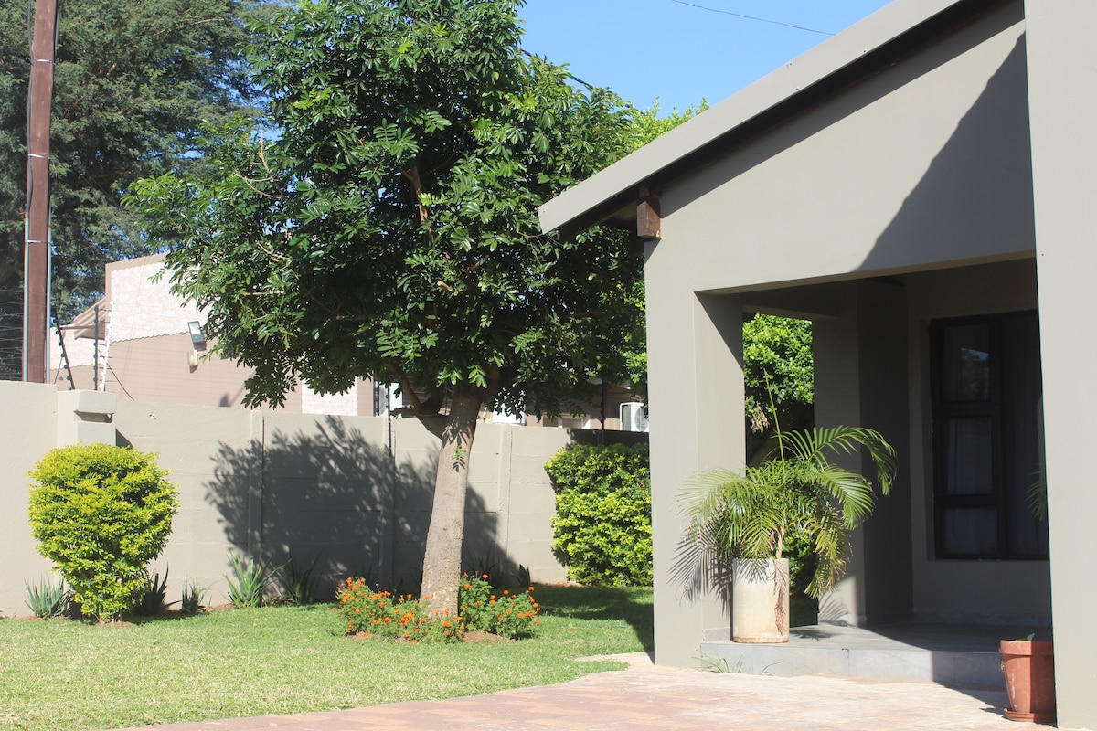A well-maintained outdoor space features a large tree providing shade alongside neatly trimmed bushes and vibrant flowers. The entrance of the building is framed by greenery, with a small planter placed at the base of the steps leading to the door.