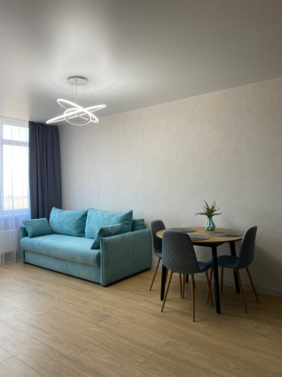 A light-filled living area features a soft teal sofa and a round dining table surrounded by four sleek chairs. A modern light fixture hangs overhead, and a small vase with fresh flowers is placed on the table. Large windows allow natural light to enhance the space.