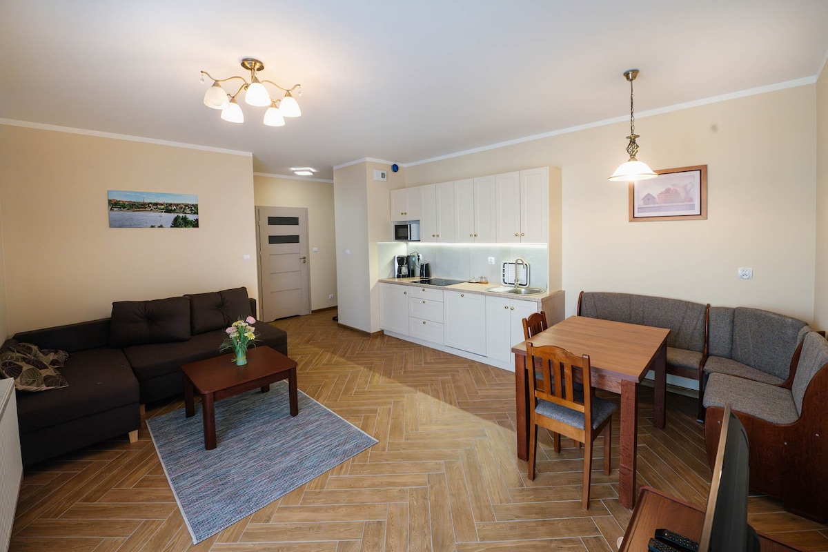 An open-plan living area features a comfortable dark sofa and a wooden dining table set with four chairs. A kitchen area with modern appliances is visible, along with a decorative rug on the beige-hued floor. Soft lighting enhances the welcoming ambiance.