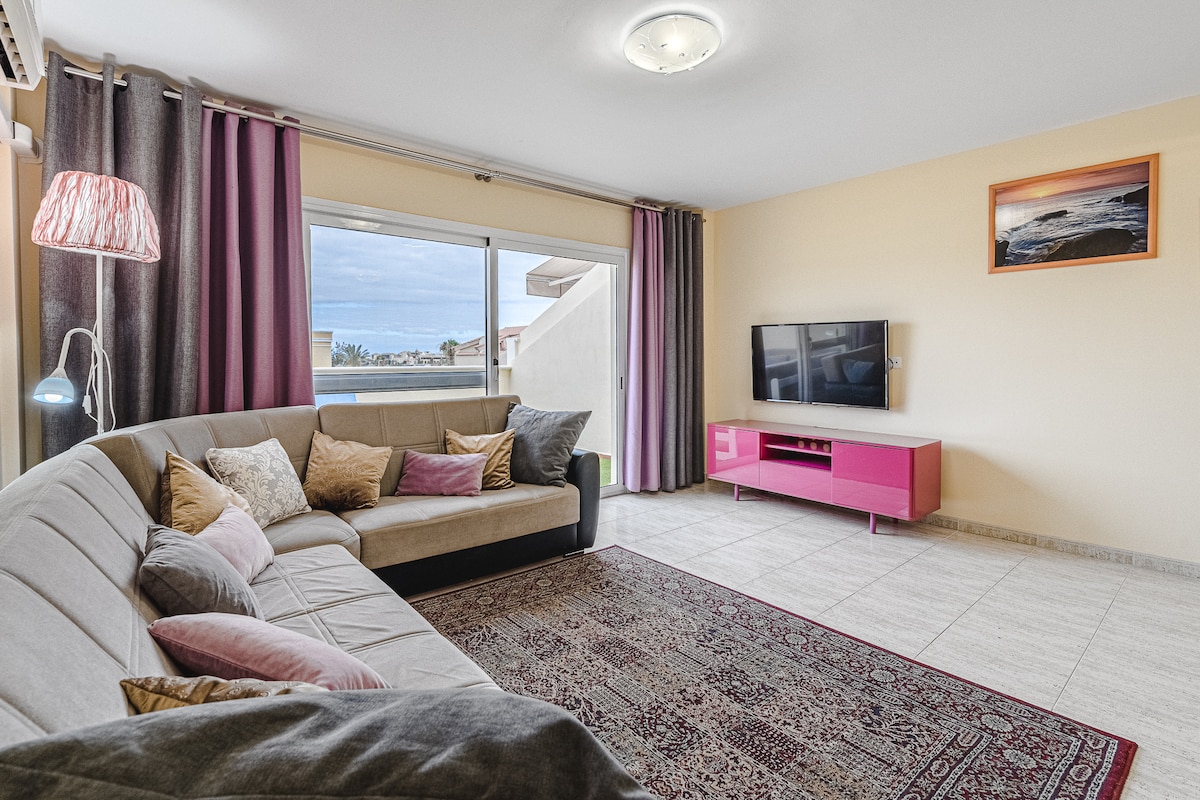 A spacious living area features a large sectional sofa adorned with decorative pillows. A flat-screen TV is mounted on the wall opposite the sofa, and large windows let in natural light, revealing views beyond. A stylish pink entertainment unit complements the decor.