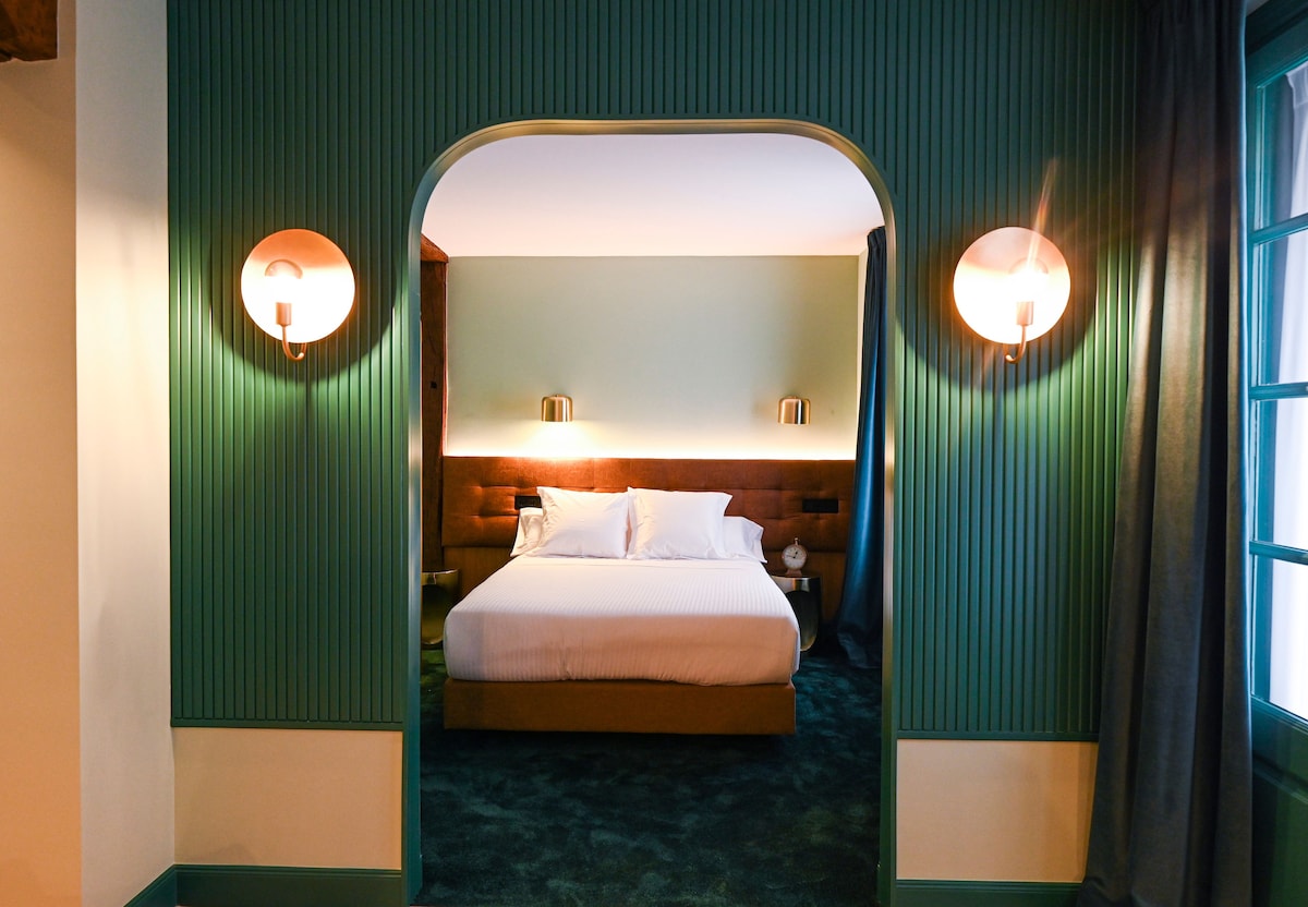 A tranquil bedroom is framed by an arched entrance, featuring a spacious bed against a soft green wall. Gentle lighting is provided by wall sconces, and thick curtains add a touch of warmth. The flooring is adorned with plush carpet in a coordinating hue.