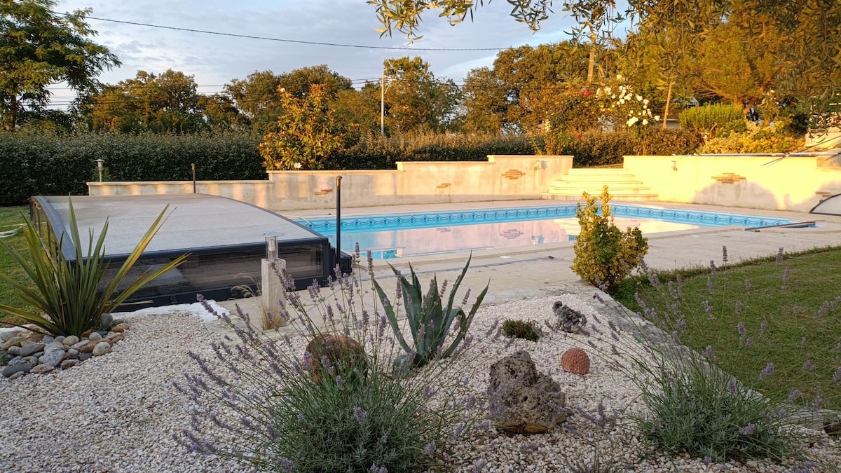 An outdoor swimming pool is surrounded by a well-maintained landscape featuring smooth pebbles and various plants. The pool area is bordered by sunlit tiles, and natural greenery offers a serene backdrop. A covered terrace can be seen in the distance, enhancing the relaxed outdoor environment.