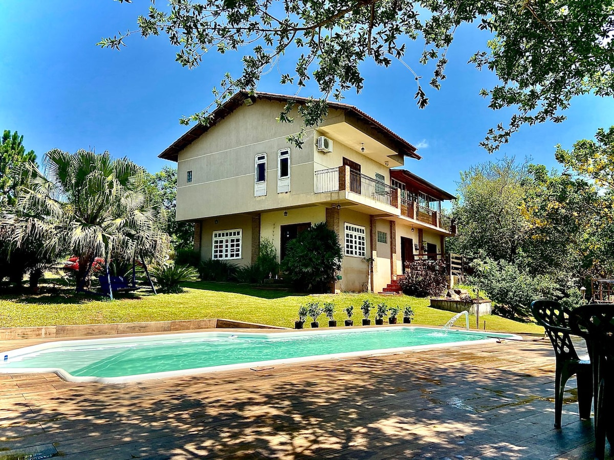 A spacious house is set against a backdrop of lush greenery. A sparkling pool is visible in the foreground, accompanied by a patio area featuring several black chairs. The upper floor includes a balcony overlooking the serene landscape.
