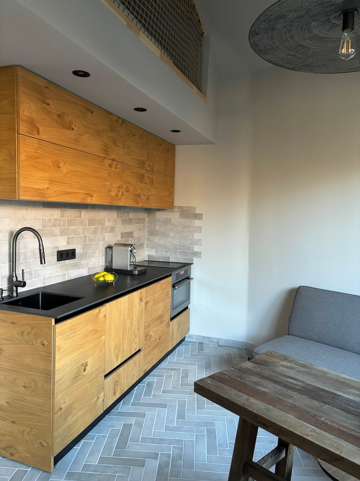 A modern kitchen area features wooden cabinetry with a sleek black countertop. Stainless steel appliances, including a kettle and an oven, are visible. A cozy seating area with a gray sofa complements a rustic wooden table, and herringbone patterned tiles enhance the space.