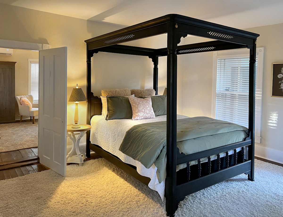 A queen-size canopy bed occupies the center of a softly lit room, featuring a light-colored comforter and decorative pillows. A bedside lamp casts a warm glow, complemented by neutral tones throughout. The space is enhanced by a plush rug and natural light filtering through window blinds.