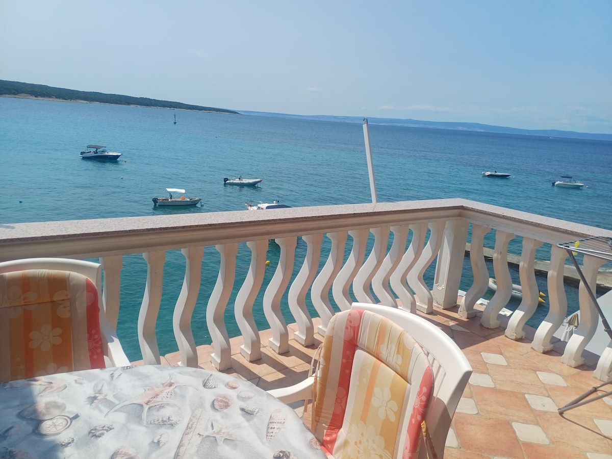 A balcony overlooks the serene blue sea, with multiple boats anchored in the water. A round table is set with patterned tablecloths and two chairs, providing an inviting space for relaxation and enjoying the coastal views.