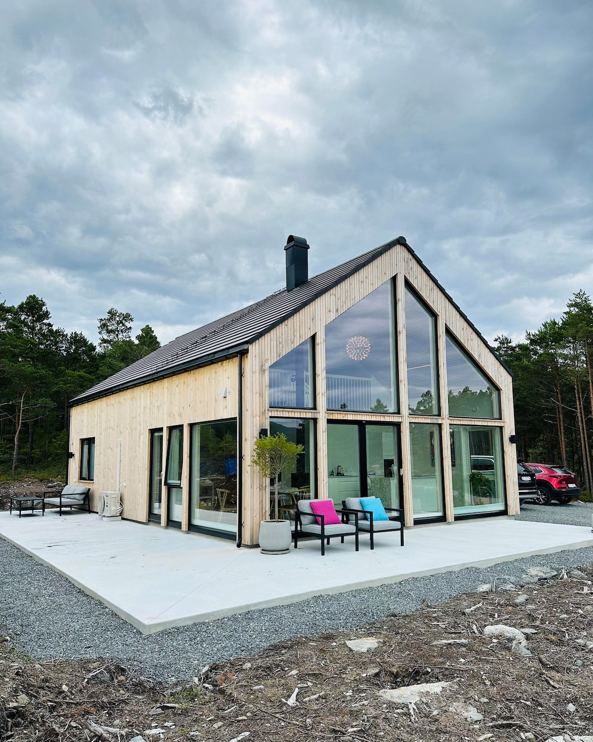 The modern cabin features a wooden exterior with large glass panels, allowing ample natural light. A spacious outdoor patio is visible, furnished with vibrant seating. Surrounding trees add a touch of nature, complementing the contemporary design of the structure.