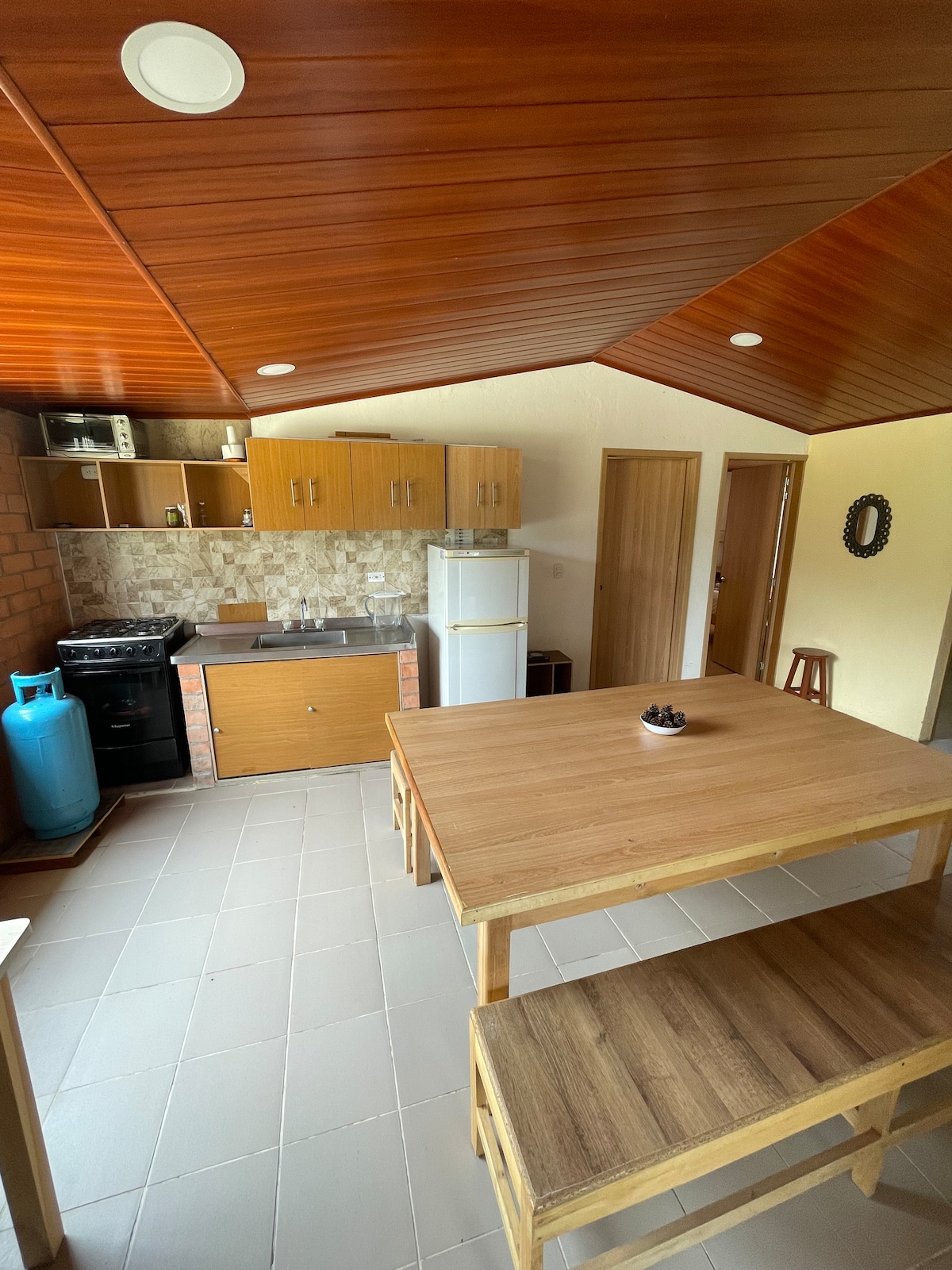 The kitchen area showcases wooden cabinets and a refrigerator, complemented by a gas stove. A wooden dining table and benches provide a casual eating space. The ceiling is accented with wooden beams, and the floor is laid with neutral tiles, promoting a functional and inviting atmosphere.
