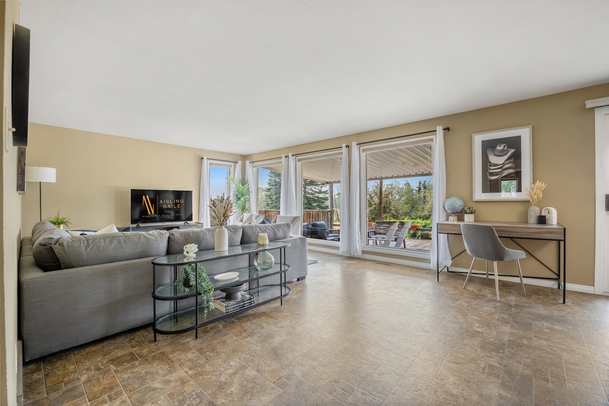 The main floor living room features a spacious sectional sofa, a coffee table, and a desk with a comfortable chair. Large sliding glass doors provide access to the oversized deck, while natural light illuminates the neutral-toned walls and tasteful decor.