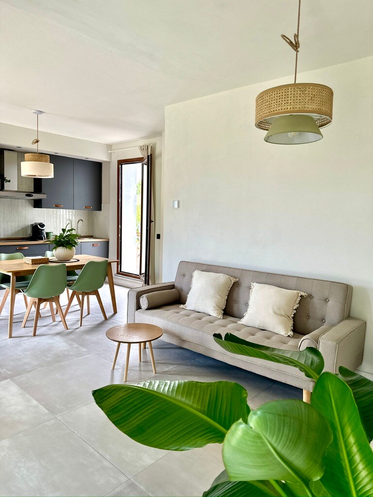 A modern living space is showcased with a neutral color palette. A comfortable sofa with pillows is positioned near a small side table. A dining table with chairs is visible in the background, accompanied by a window that allows natural light to fill the room.