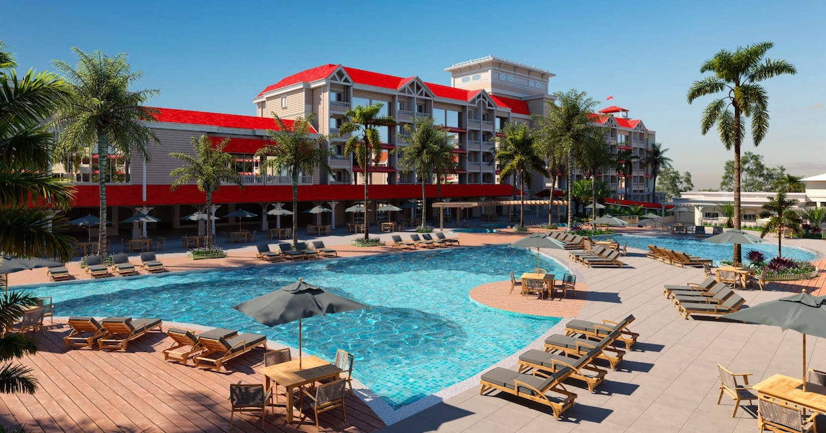 A large, inviting pool area is surrounded by lounge chairs and umbrellas. The resort features a multi-level design with red roofs and palm trees, creating a vibrant outdoor atmosphere. Sunlight reflects off the water, enhancing the tranquil setting for relaxation.