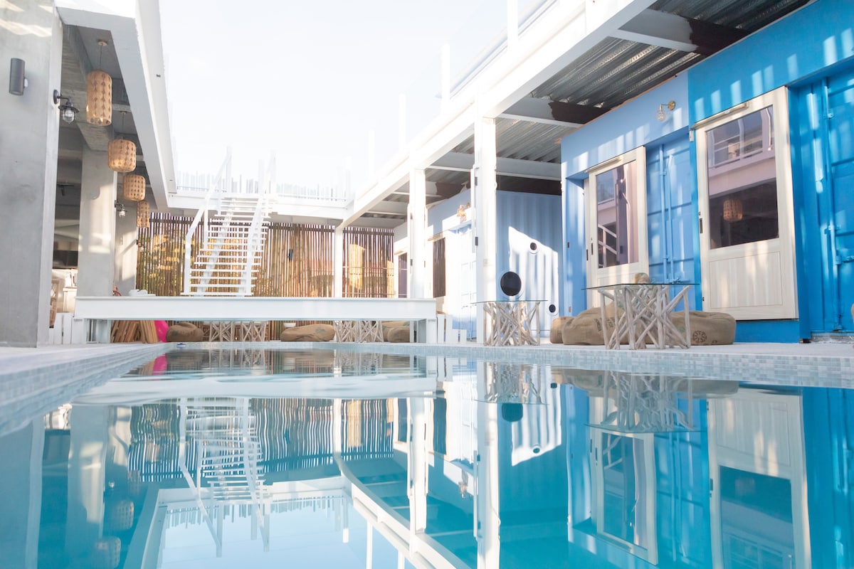 A swimming pool reflects the surrounding structures, which exhibit a mix of industrial and coastal designs. Light wooden furnishings and lounge areas provide a relaxed atmosphere. The upper level is accessible by a staircase, while vibrant blue walls complement the overall aesthetic.