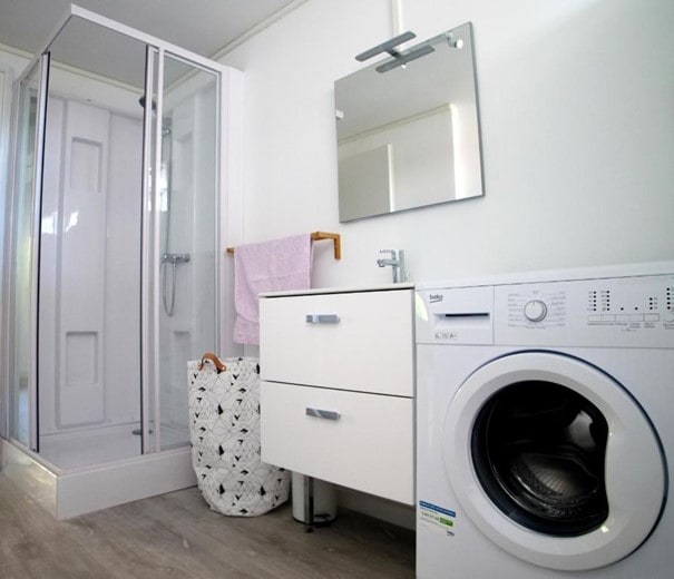 A functional bathroom features a glass-enclosed shower unit, a modern sink with a mirror above, and a washing machine. A white cabinet offers storage, while a woven laundry basket adds texture to the space. Light-colored walls contribute to a clean and uncluttered appearance.