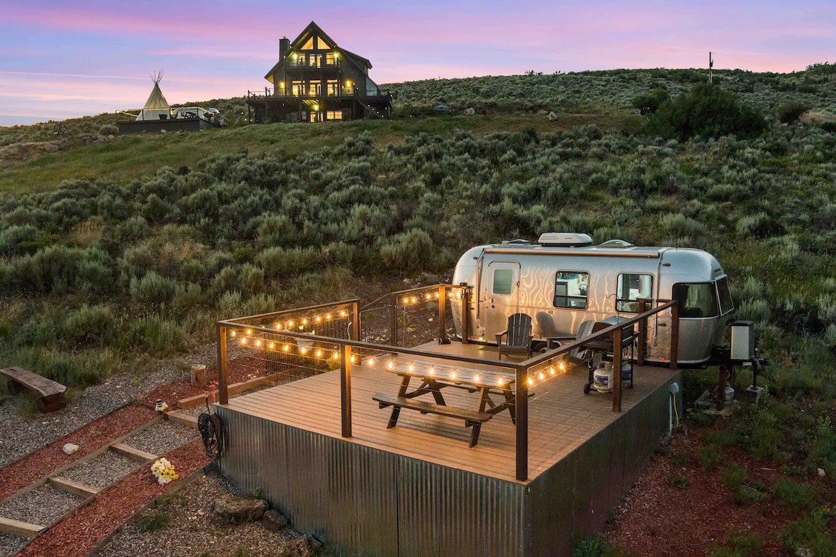 A modern Airstream is positioned on a spacious wooden deck, adorned with string lights that create a warm ambiance. Surrounding the area is lush green shrubbery under a vibrant sky at dusk. A large lodge in the background complements the serene mountain landscape.