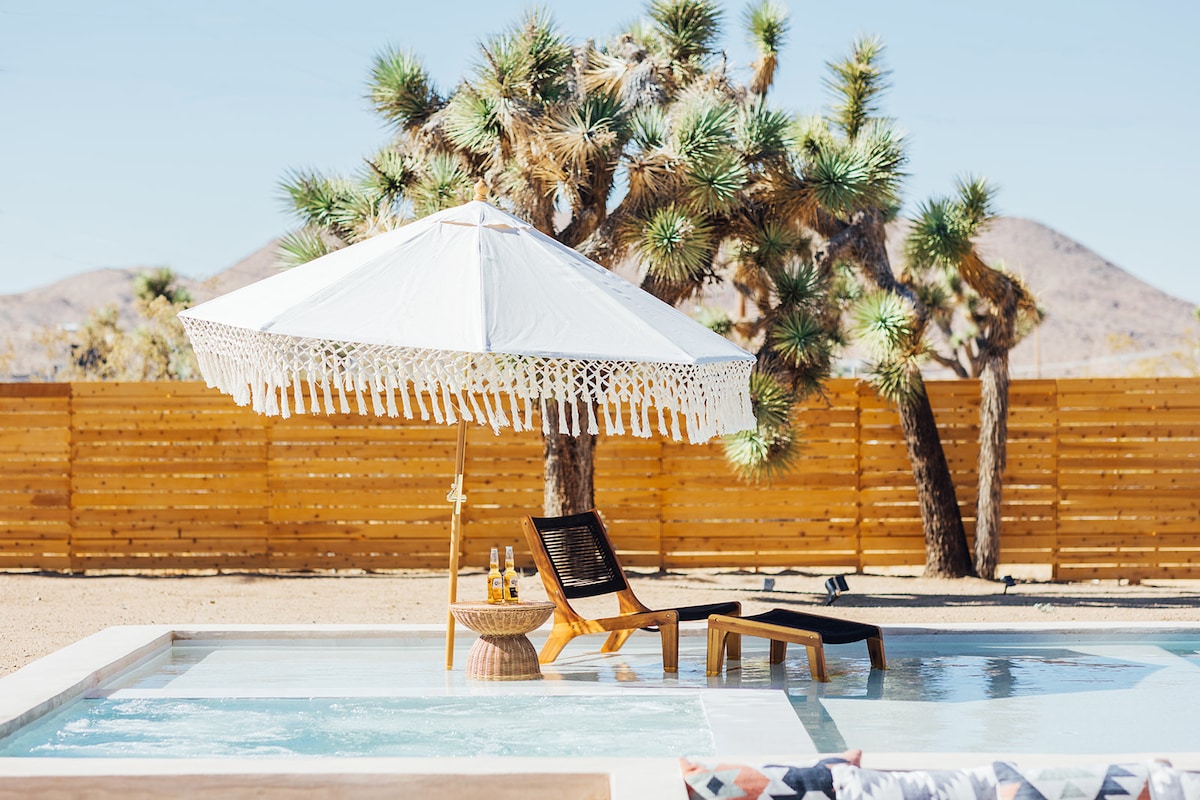 A resort-style swimming pool is featured, complete with an oversized tanning shelf. A large white umbrella provides shade over dark wooden lounge chairs. Joshua trees are visible in the background, alongside a wooden fence that encloses the backyard.