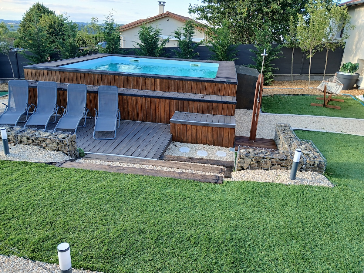 An outdoor space features a shallow, rectangular pool surrounded by wooden decking. Six gray lounge chairs are arranged nearby on lush green grass. A stone wall segments the area, and LED lighting lines the pathway, providing illumination for evening relaxation.