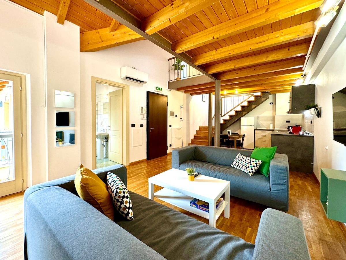 A modern living area features two spacious sofas adorned with colorful cushions. A white coffee table sits in the center, while warm wooden beams span the ceiling. The open space connects to a staircase leading to the upper level, and the kitchen is visible in the background.
