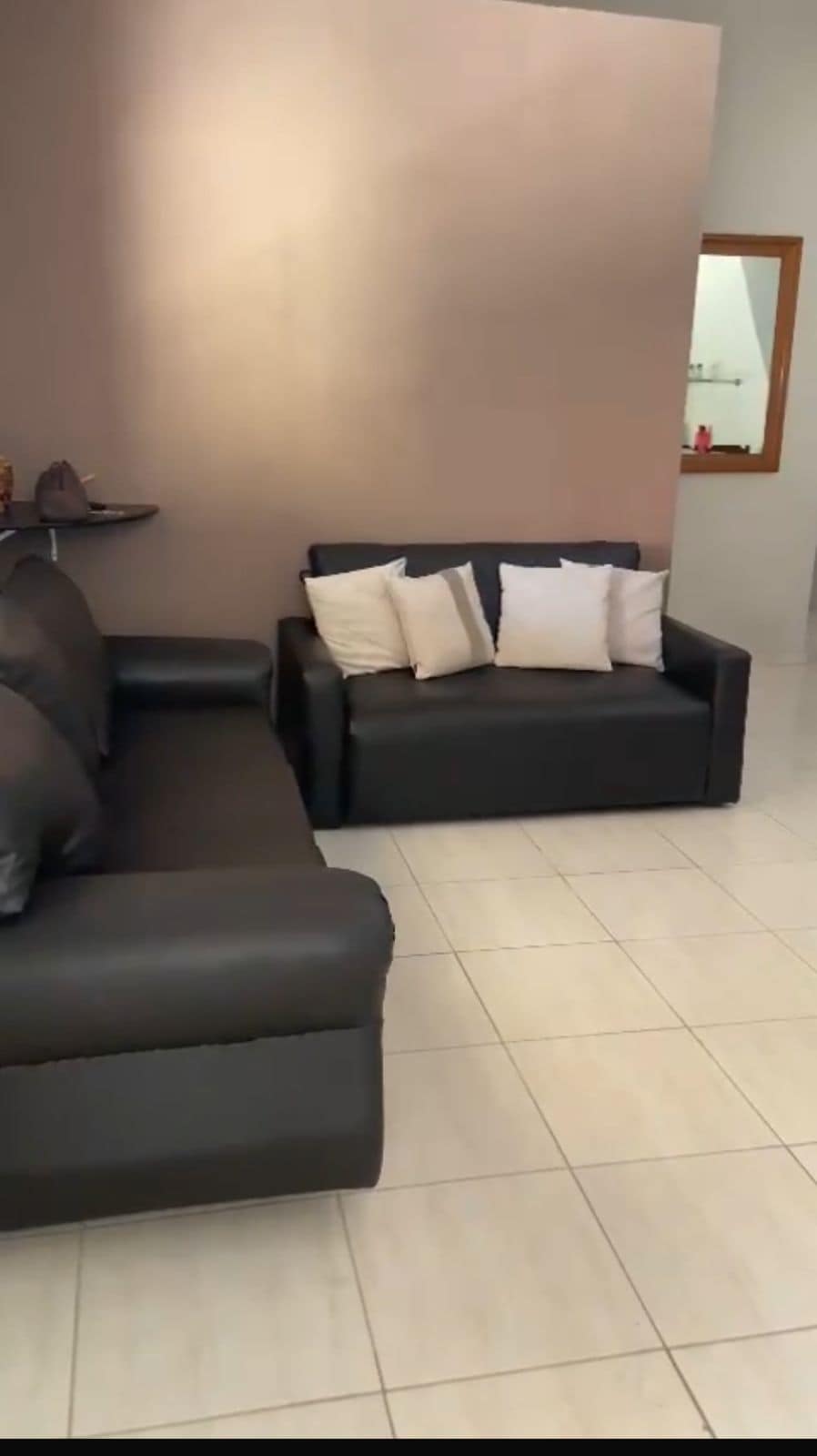 A living area features two black sofas, one larger with armrests and a smaller loveseat. Decorative cushions are arranged on the smaller sofa. Light tiles cover the floor, while a neutral wall provides a warm backdrop, creating a spacious atmosphere.