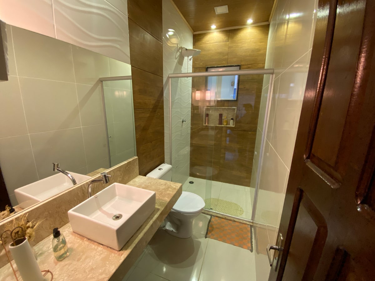 The bathroom features a dual sink vanity with square basins and a modern countertop. A glass-enclosed shower is visible, alongside a compact toilet. Warm wood accents create a calming environment, complemented by soft lighting overhead.