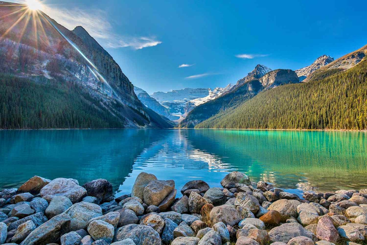 A scenic view of Lake Louise is presented, with tranquil turquoise waters reflecting the surrounding mountains and lush evergreen trees. Rocky shorelines frame the lake, and the sun casts a warm glow over the landscape, enhancing the natural beauty of the area.