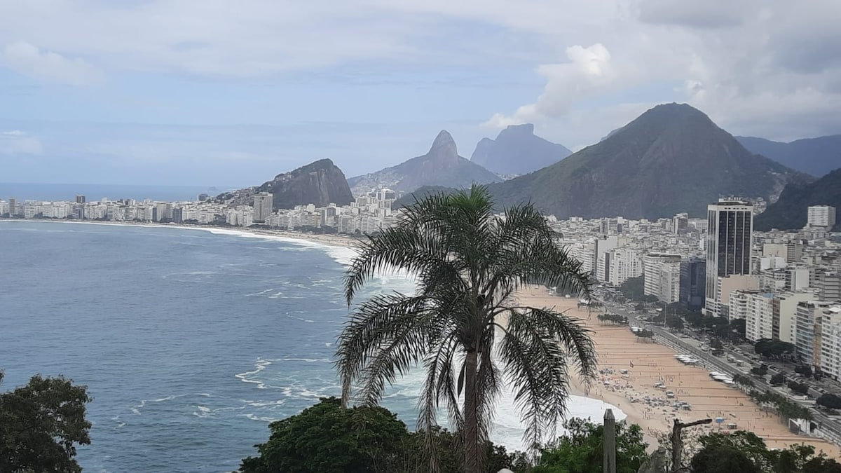 A panoramic view captures the coastline of Copacabana Beach, with waves gently lapping the shore. The image features distant mountains and a beach lined with umbrellas. Palm trees in the foreground add a touch of green to the vibrant landscape under a partly cloudy sky.