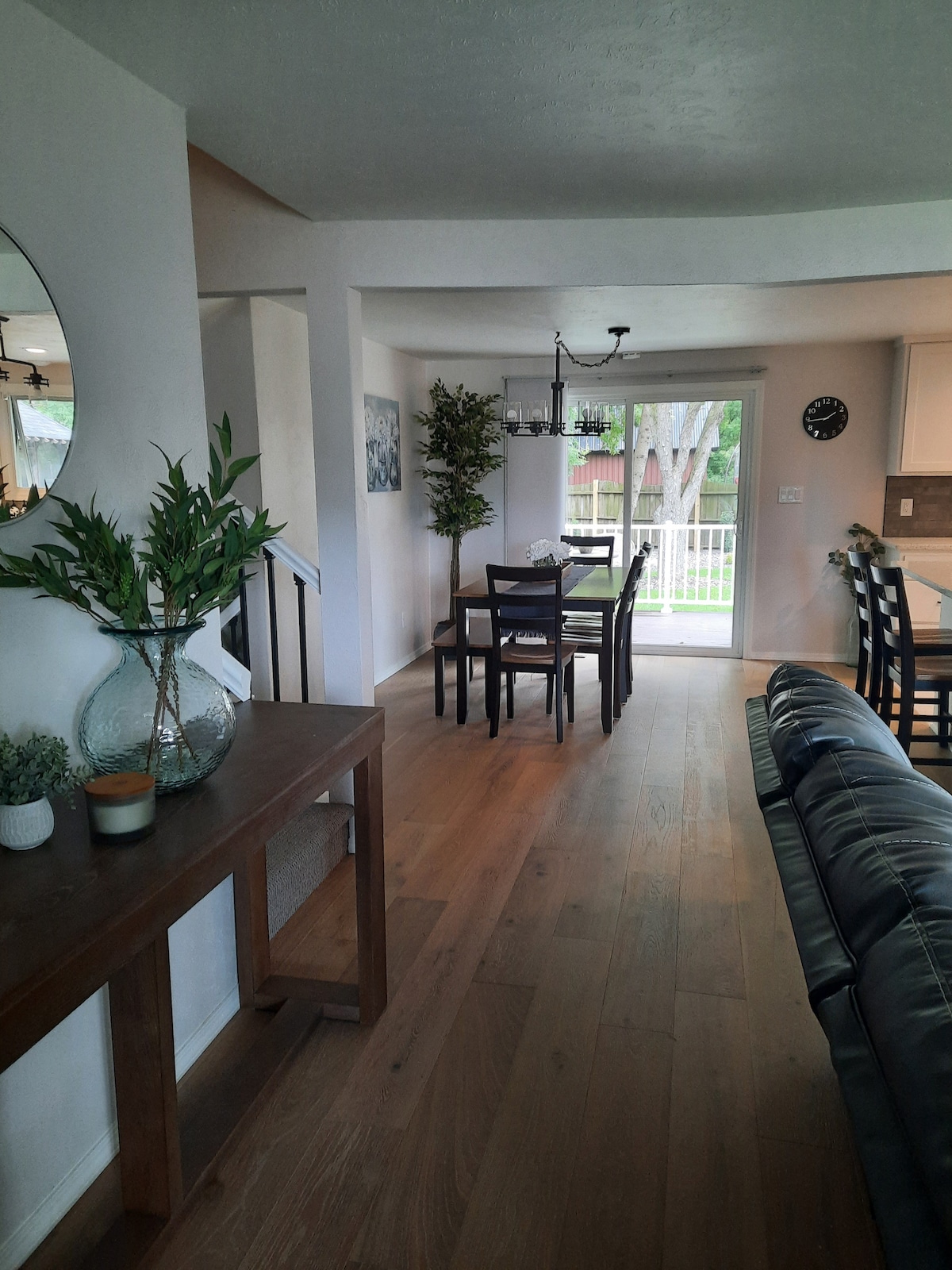 A dining area is situated next to large sliding doors that provide natural light and views of the backyard. A wooden table with six chairs is positioned centrally. A console table holding decorative items enhances the space, framed by greenery and soft color tones.