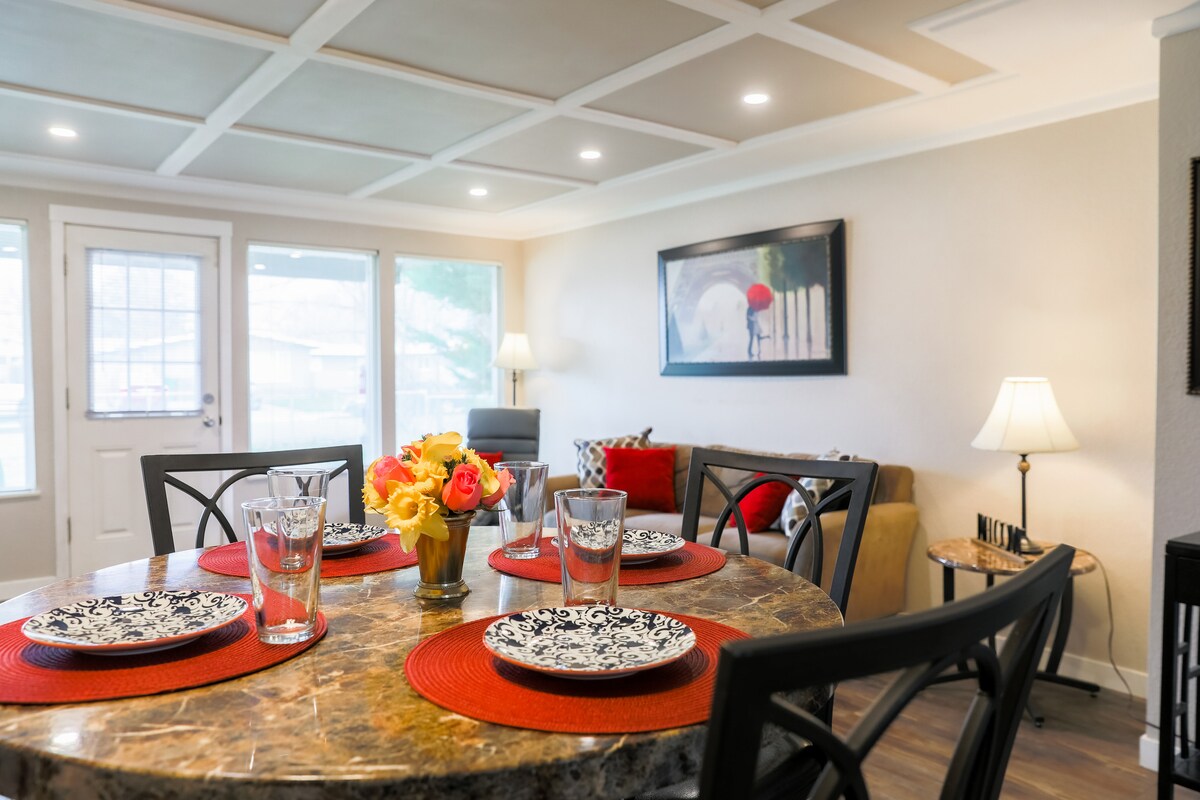 A dining area is highlighted with a round table set for four, featuring decorative plates and glasses. A small floral centerpiece adds color. Natural light fills the space, with a cozy seating area visible in the background.