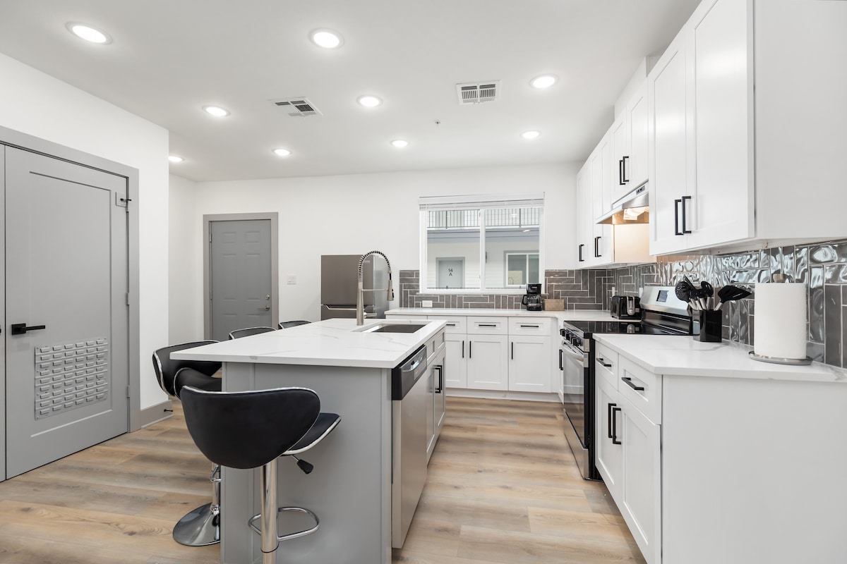 A modern kitchen is equipped with sleek white cabinetry and a large island featuring a built-in sink. Stainless steel appliances are present, including a microwave and an oven. A window allows natural light to illuminate the space, complemented by recessed lighting overhead.