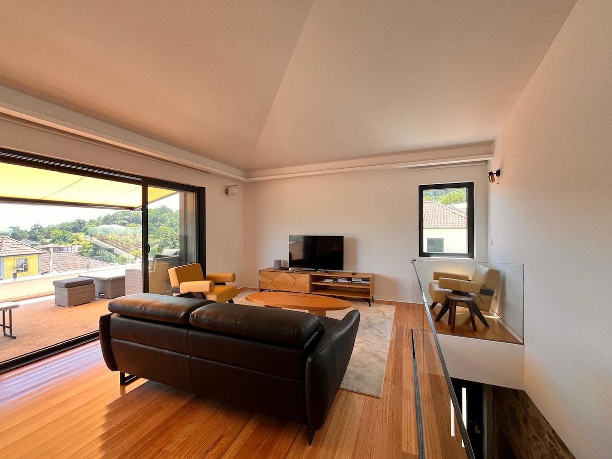 A spacious living area features large windows that invite natural light and provide views of the outdoor terrace. Contemporary furniture includes a dark leather sofa and modern armchairs. A wooden entertainment unit holds a flat-screen television, and a light rug is positioned on the warm wooden floor.