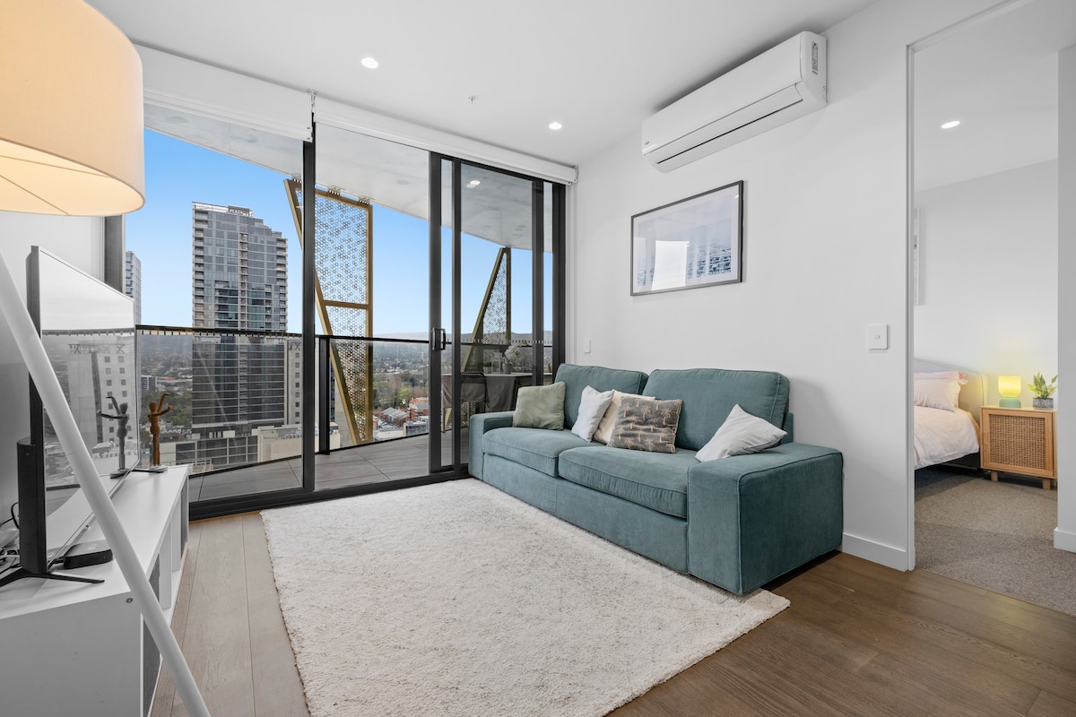 A modern living area features a plush teal couch positioned on a light area rug. Large glass doors open to a balcony, offering views of the city skyline. A TV stand holds a television, while artwork decorates the walls, contributing to an inviting ambiance.