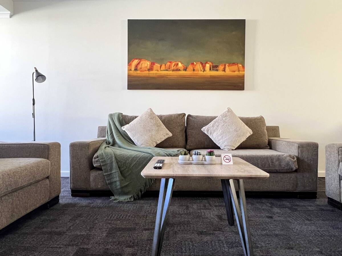 A comfortable living area features a sofa adorned with decorative cushions, complemented by a textured throw. A coffee table with a small plant and a remote control sits in front, and a large artwork depicting a landscape is displayed on the wall behind the seating.