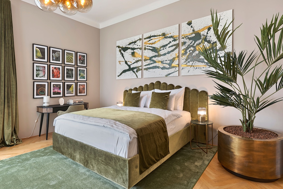 A spacious bedroom features a plush green bed with elegant headboard and complementary bedding. Artwork is displayed on the wall above a bedside table, while a large plant and decorative lamp add character. Natural light filters through a nearby window covered by a rich curtain.