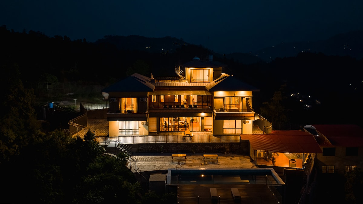 The exterior of a modern villa is illuminated at night, showcasing its spacious design and multiple levels. Warm lights reflect on the surrounding landscape, creating a serene atmosphere. A clear view of the patio and pool area can be seen below, set against the darkened backdrop of the hills.