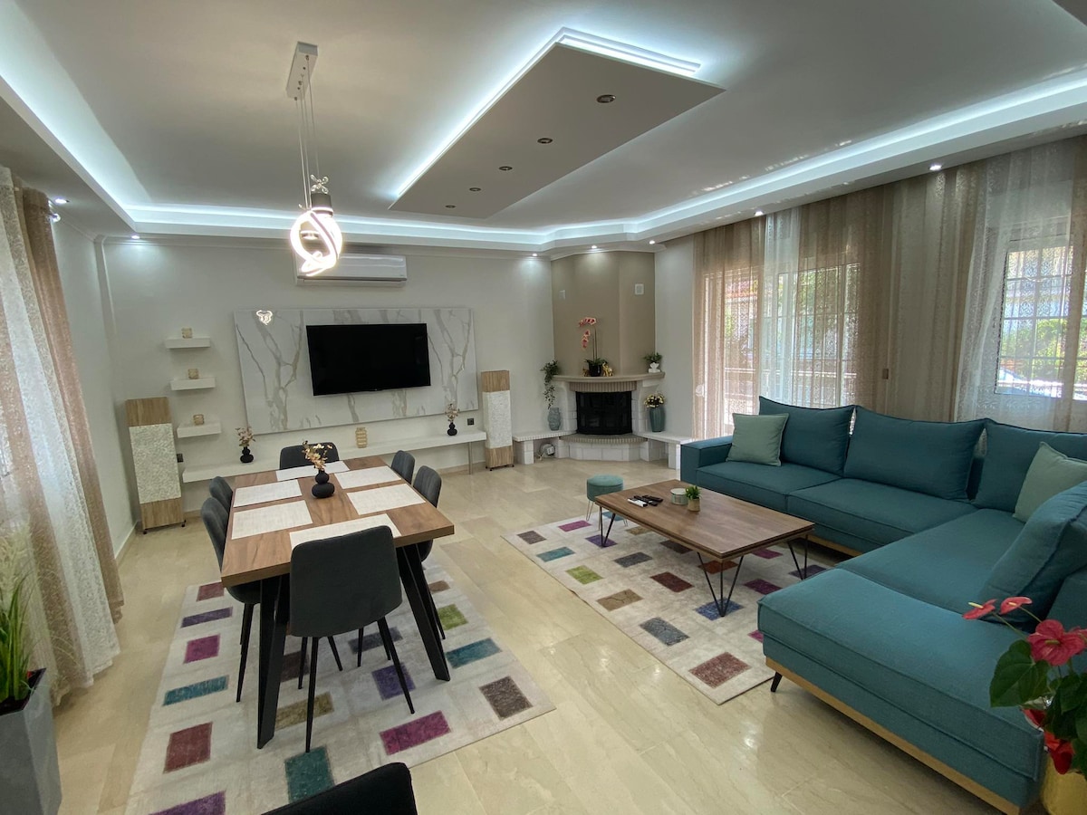 A spacious living area is shown, featuring a turquoise sectional sofa and a wooden dining table set for six. Bright natural light filters through sheer curtains, and decorative elements, including a flat-screen TV and green plants, are placed throughout the room.