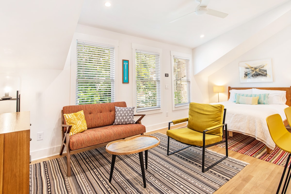 A light-filled seating area includes a brown sofa with decorative pillows and an accent chair in a vibrant yellow hue. A wooden coffee table sits on a patterned rug, and a spacious bed is positioned nearby, illuminated by natural light from the windows.