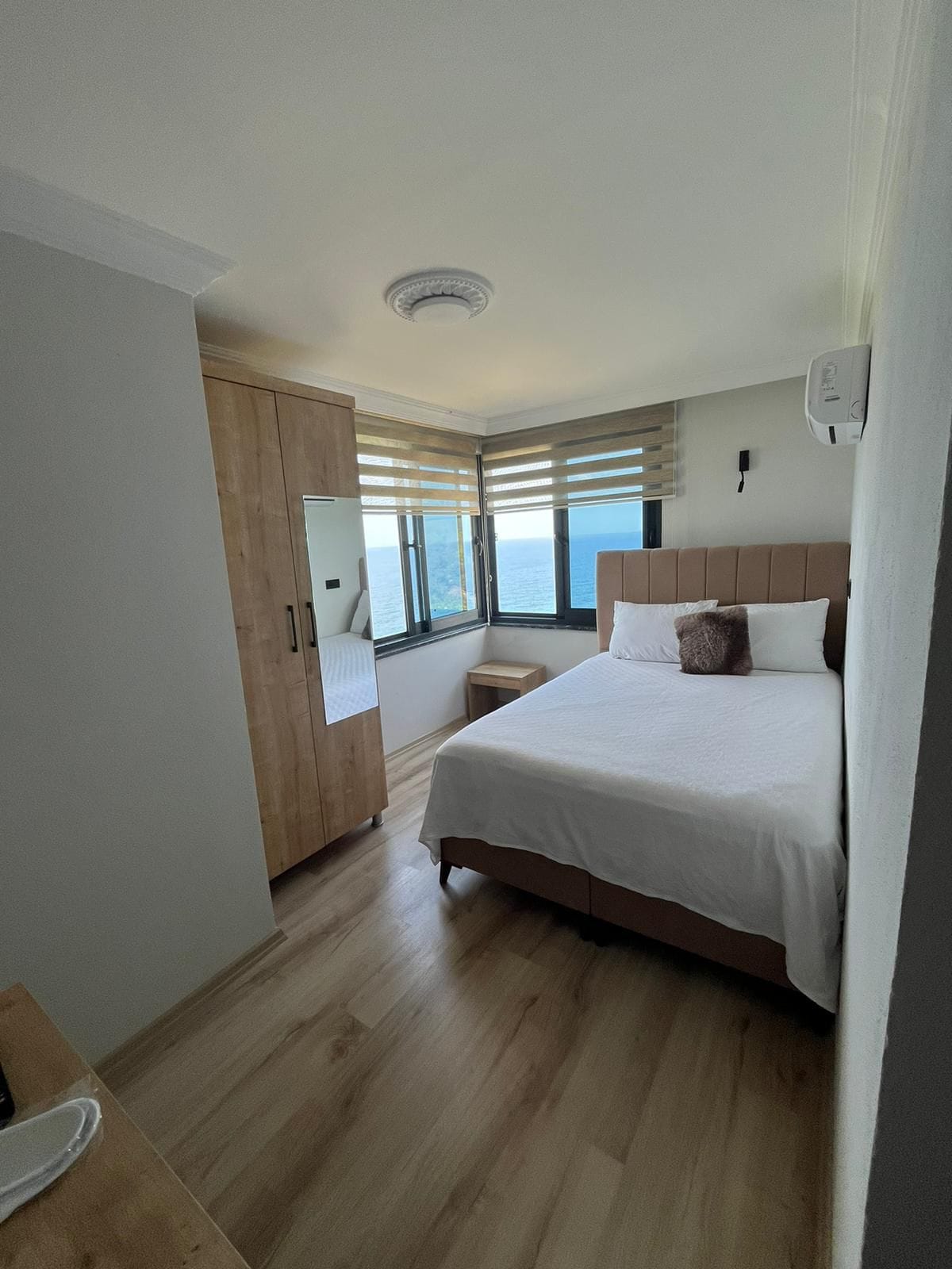 A bright and airy bedroom is highlighted by large windows offering views of the ocean. A spacious bed with crisp white linens is complemented by a neutral headboard. A wooden wardrobe and bedside table add functionality to the serene space.
