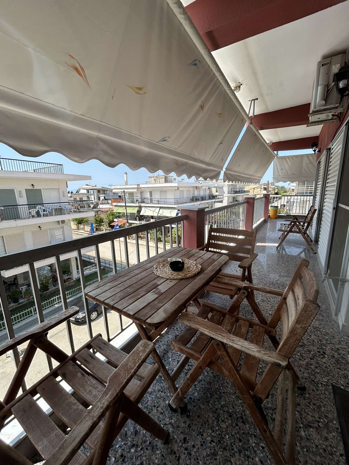 A shaded balcony is seen with wooden furniture, including a table and chairs. The area is adorned with a round woven placemat and a small black item placed at the center of the table. Nearby buildings and a street are visible beyond the railing.