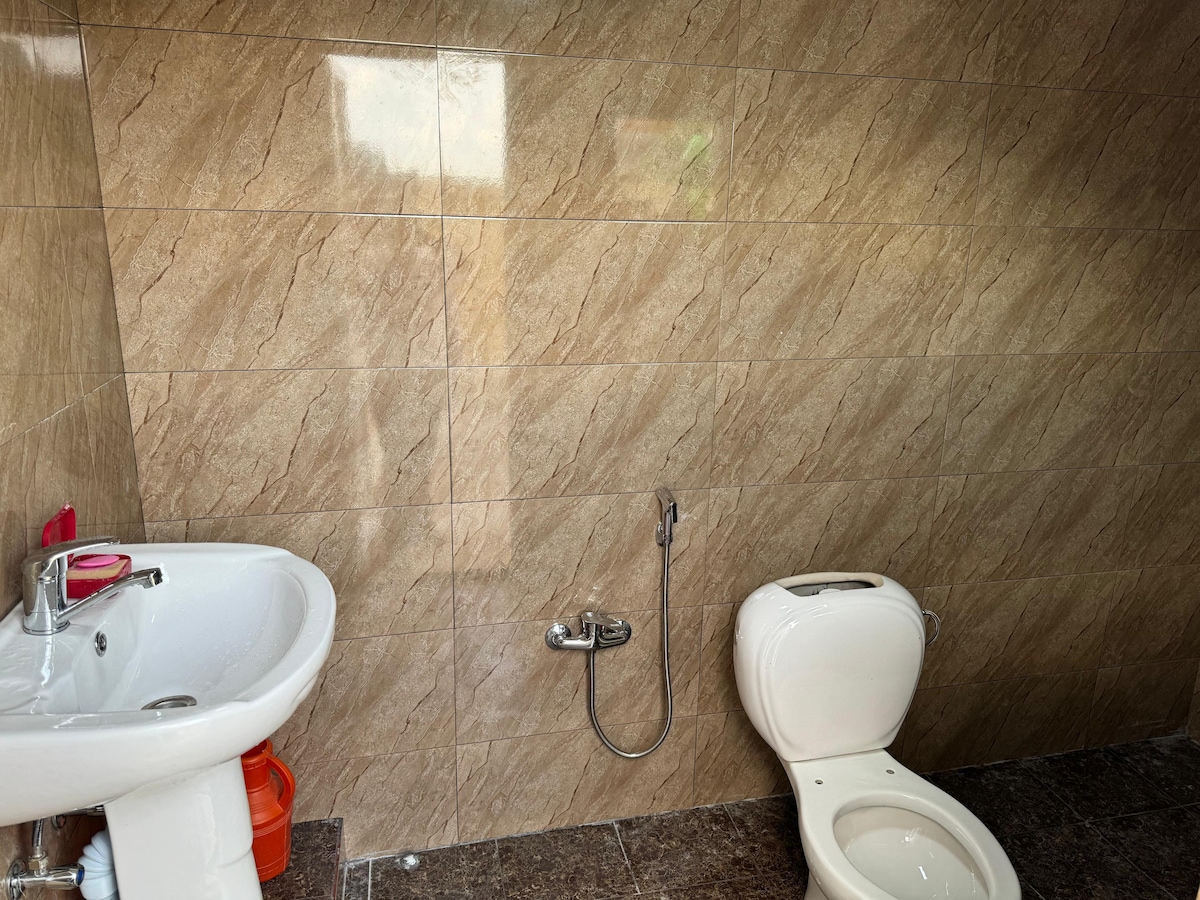 A clean and functional bathroom is presented, featuring a white toilet and a wall-mounted sink. The walls are covered in light brown tiles, providing a modern look, while a shower fixture is visible next to the sink.
