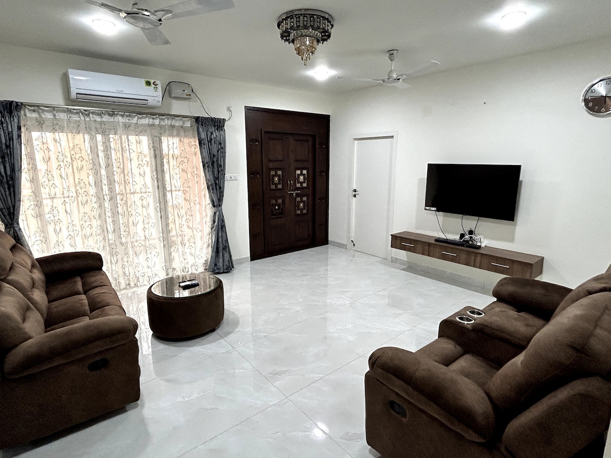 The living hall features two recliner sofas in rich brown upholstery, complemented by a circular coffee table. A 50-inch television is mounted on the wall opposite the seating area, while sheer curtains frame the windows, allowing natural light to filter softly into the space.