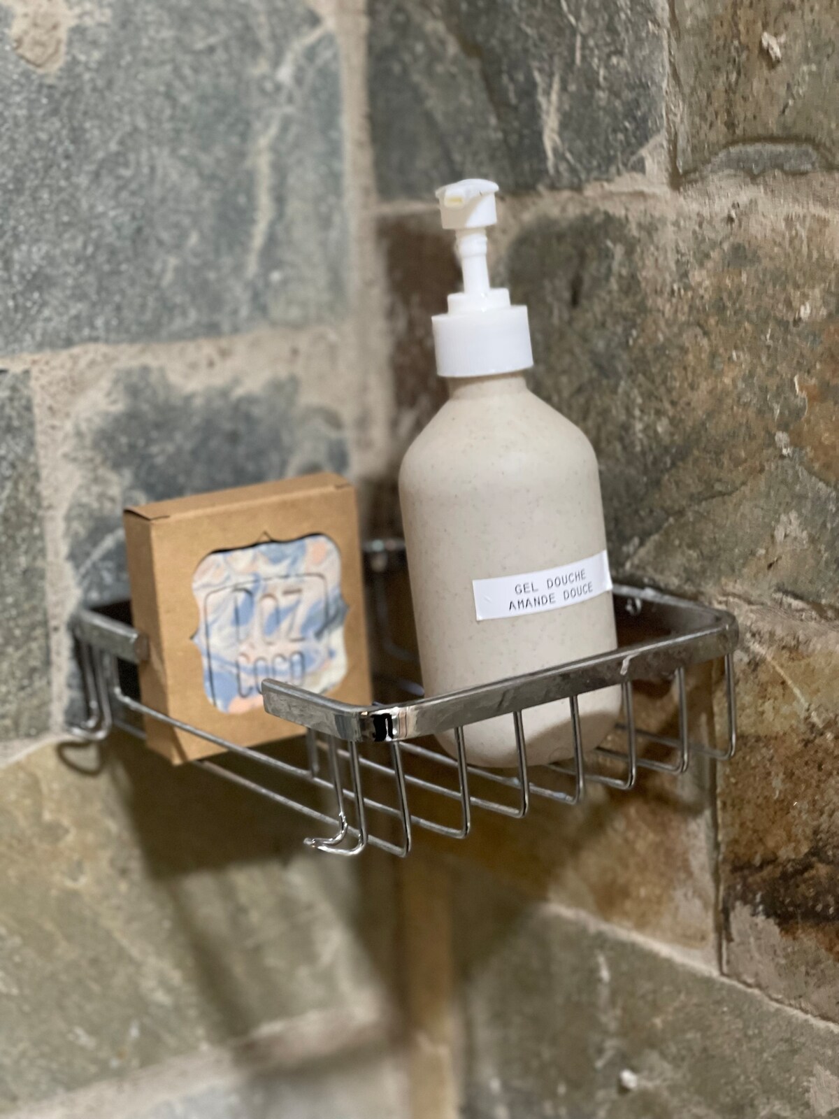 A silver shower caddy is attached to a textured stone wall. It holds a pump bottle of body wash and a small cardboard box labeled 'soap.' The neutral tones of the products blend harmoniously with the natural stone background.