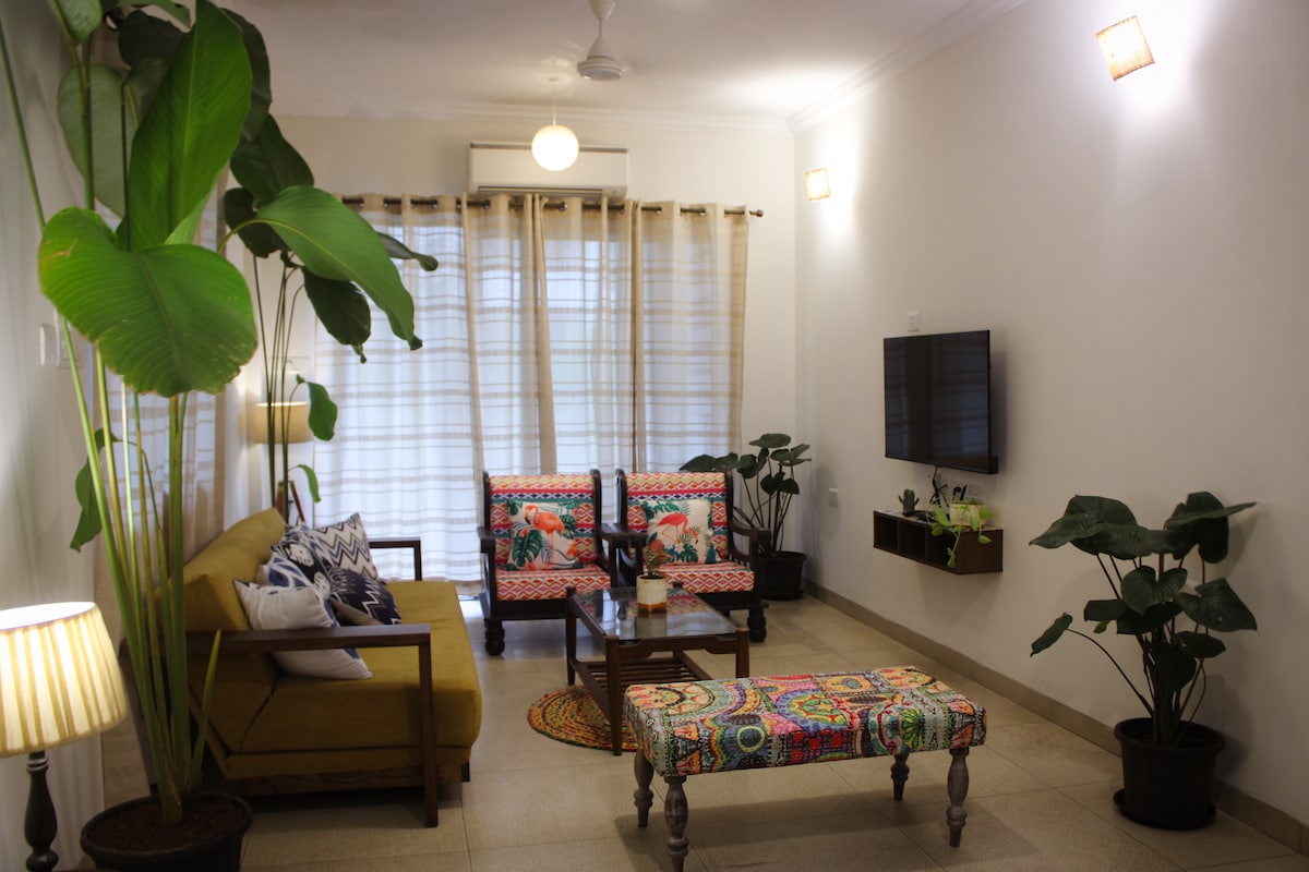 A bright living area is depicted with a comfortable sofa and colorful patterned armchairs. A coffee table sits at the center, surrounded by potted plants. Natural light enters through sheer curtains, enhancing the space's inviting ambiance, while a wall-mounted television is visible.