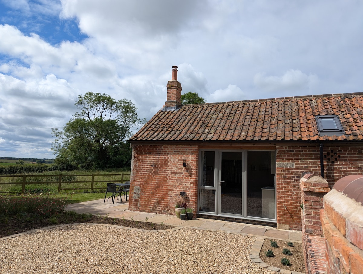 The exterior of a brick building features a sloped roof with a skylight. A patio area is visible in front, furnished with a small table and two chairs. Lush greenery surrounds the property, providing a serene countryside atmosphere.
