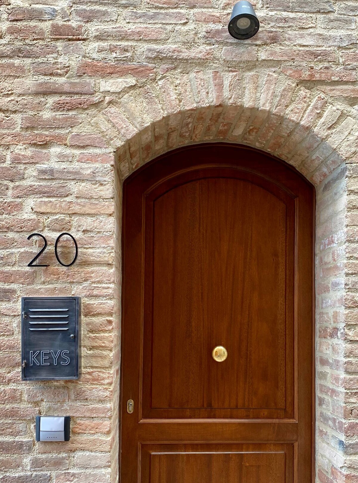 An arched wooden door is framed by a rustic brick wall, featuring a circular brass doorknob. A rectangular mailbox with the word 'KEYS' is mounted beside the entrance, which is illuminated by a wall light above.